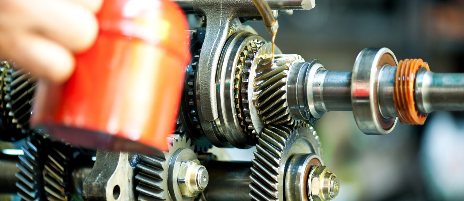 A close-up view of a complex gear system being lubricated with oil from a red canister. The image highlights the importance of proper maintenance and precision in industrial machinery, showcasing detailed gears, bearings, and moving components in a mechanical setup.